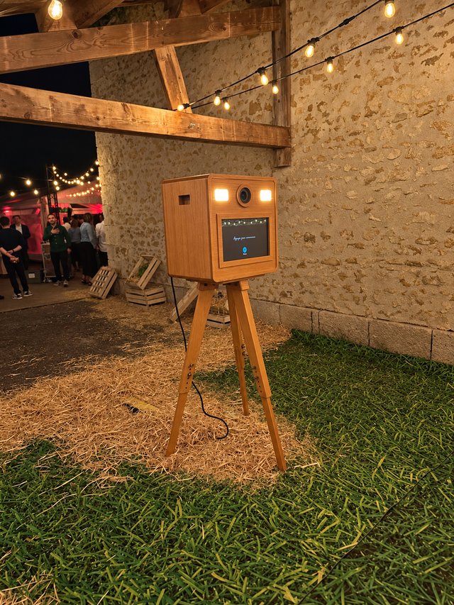 Photobooth en bois sur trépied, installé au pied d’un mur en pierre sous des guirlandes.