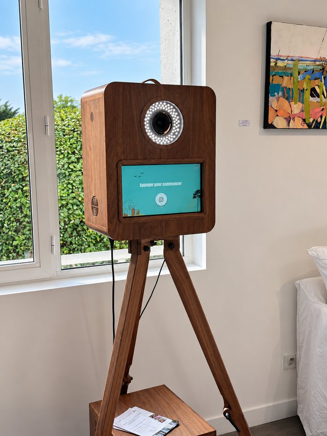 Photobooth en bois foncé sur trépied devant une fenêtre, avec écran et éclairage circulaire.